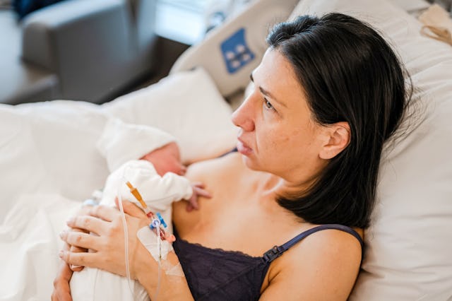 A mother tenderly holds her newborn baby while breastfeeding in a hospital bed. She looks calm and r...