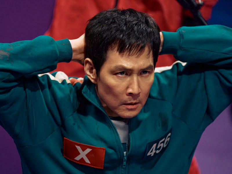 A tense man in a green jumpsuit with the number 456 stands against a purple background, gripping his...