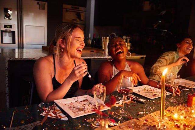 A group of three women laughing joyfully around a dinner table, with plates and glasses around them,...