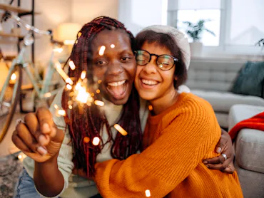Two young women joyfully embrace while holding a sparkler, surrounded by a cozy indoor setting with ...