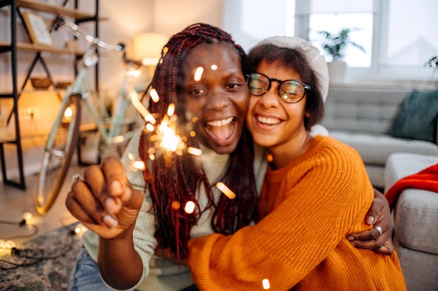 Two young women joyfully embrace while holding a sparkler, surrounded by a cozy indoor setting with ...