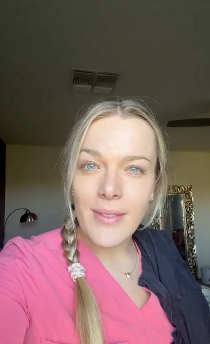 A woman with long blonde hair in a braid, wearing a pink top, smiles at the camera. She stands in a well-lit room with a decorative mirror.