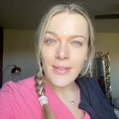 A woman with long blonde hair in a braid, wearing a pink top, smiles at the camera. She stands in a well-lit room with a decorative mirror.