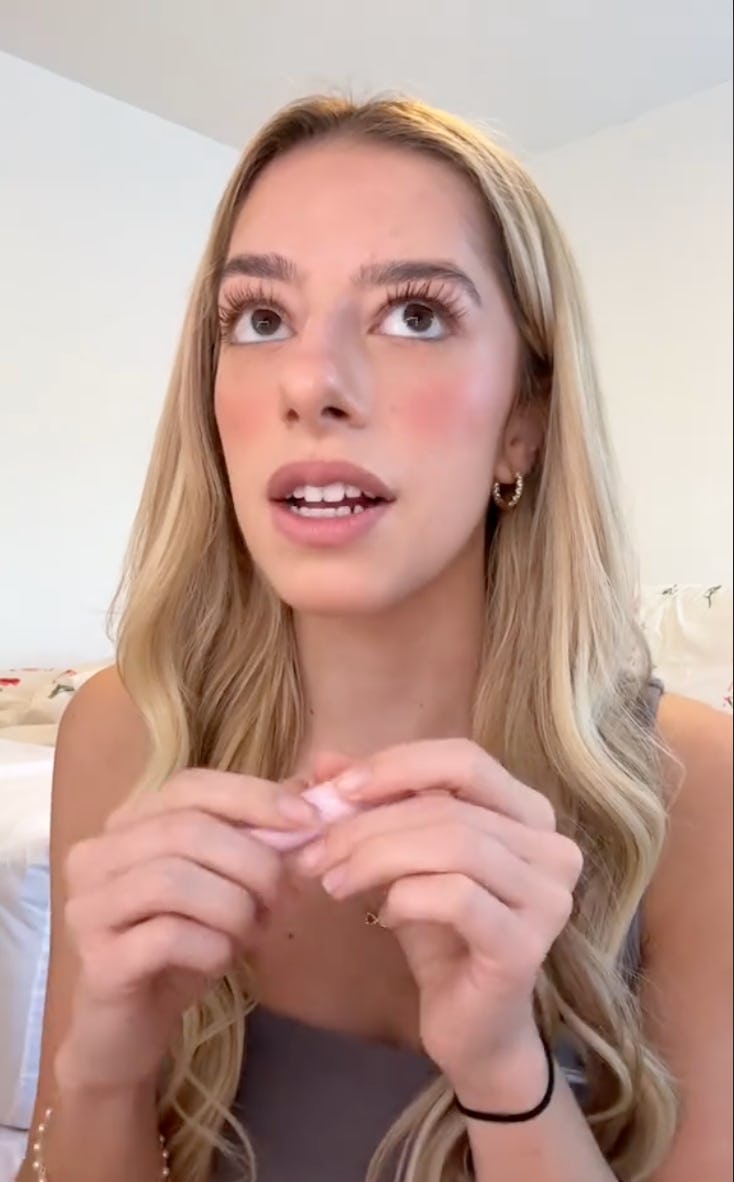 A young woman with long, wavy blonde hair is speaking and looking up. She holds a small object in her hands, wearing a simple outfit with soft makeup.