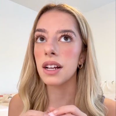 A young woman with long, wavy blonde hair is speaking and looking up. She holds a small object in her hands, wearing a simple outfit with soft makeup.