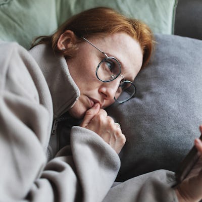 A person with red hair and glasses relaxes on a couch, holding a smartphone while resting their head on a gray pillow, appearing deep in thought.