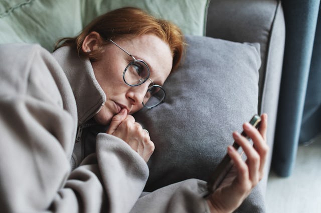 A person with red hair and glasses relaxes on a couch, holding a smartphone while resting their head...