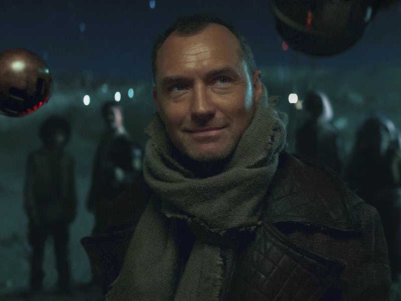 A man with a slight smile stands in a dimly lit scene, wearing a scarf and coat. Suspended metallic ...
