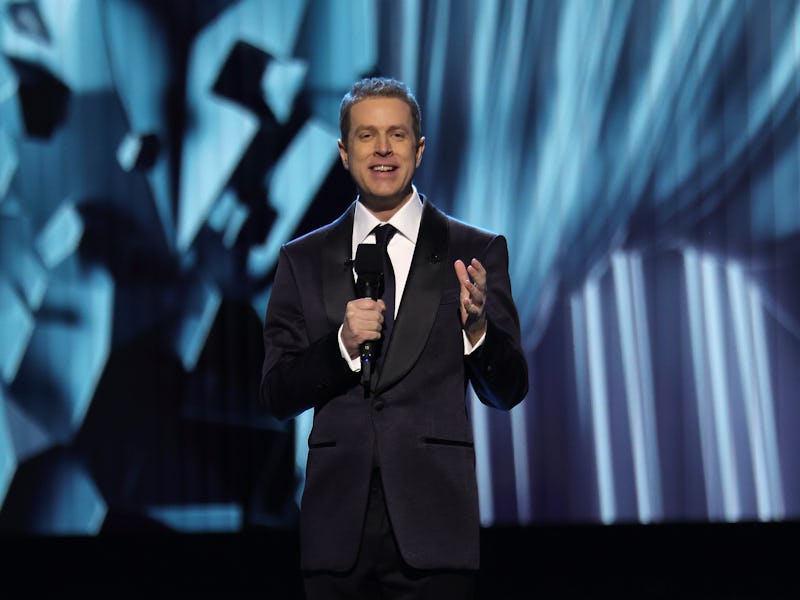 A suited speaker stands on stage, microphone in hand, against a dynamic, illuminated backdrop. The s...