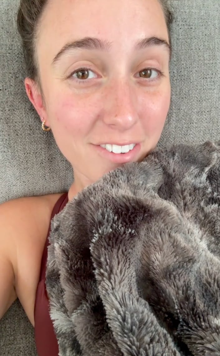 A smiling young woman lies on a gray couch, partially covered with a soft, fuzzy blanket. She has a ...