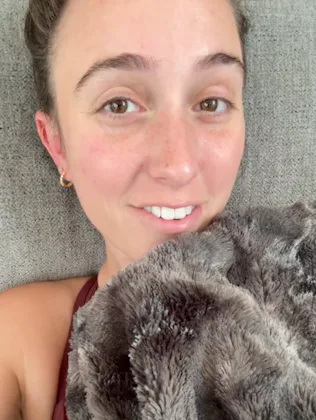 A smiling young woman lies on a gray couch, partially covered with a soft, fuzzy blanket. She has a ...