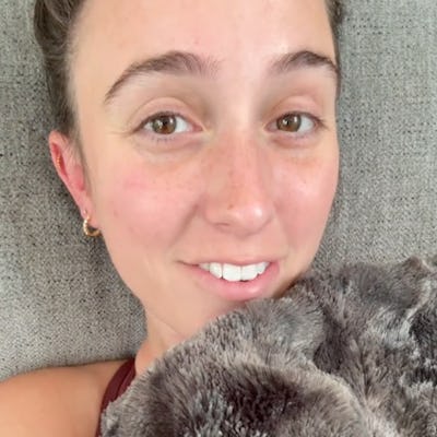 A smiling young woman lies on a gray couch, partially covered with a soft, fuzzy blanket. She has a natural look with minimal makeup and bright eyes.