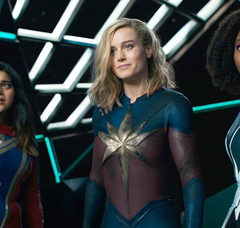 Three female superheroes stand confidently in colorful costumes against a futuristic backdrop with n...