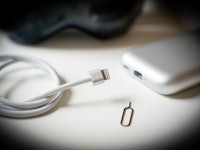 A close-up of a white charging cable and a SIM card ejector tool next to a smartphone on a white sur...