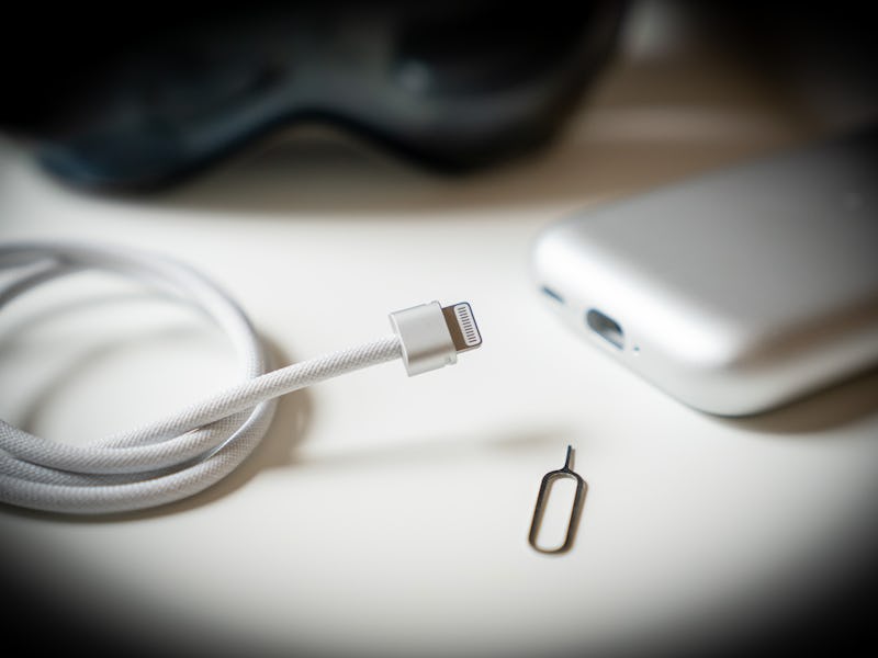 A close-up of a white charging cable and a SIM card ejector tool next to a smartphone on a white sur...