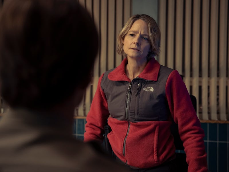 Woman in a red and gray vest looks at someone off-camera with a concerned expression, indoors near v...