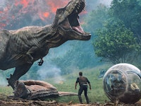 A man stands near a glass sphere vehicle while a giant dinosaur roars in a chaotic scene with smoke ...
