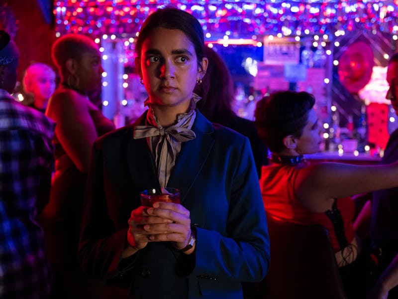 A pensive person with a drink in hand stands in a vibrant nightclub lit with colorful lights, surrou...