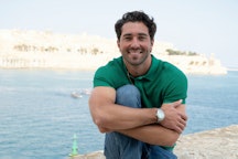 Smiling man in a green shirt sitting by the sea with a historic cityscape in the background.