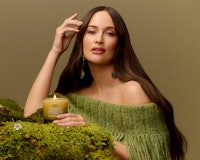 A woman in a green off-shoulder top poses with a lit candle on a bed of moss, exuding a serene ambia...