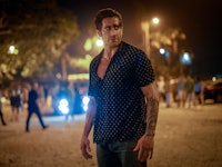 Man with a tattoo on his arm walking on a crowded beach at night, looking to the side with a serious...