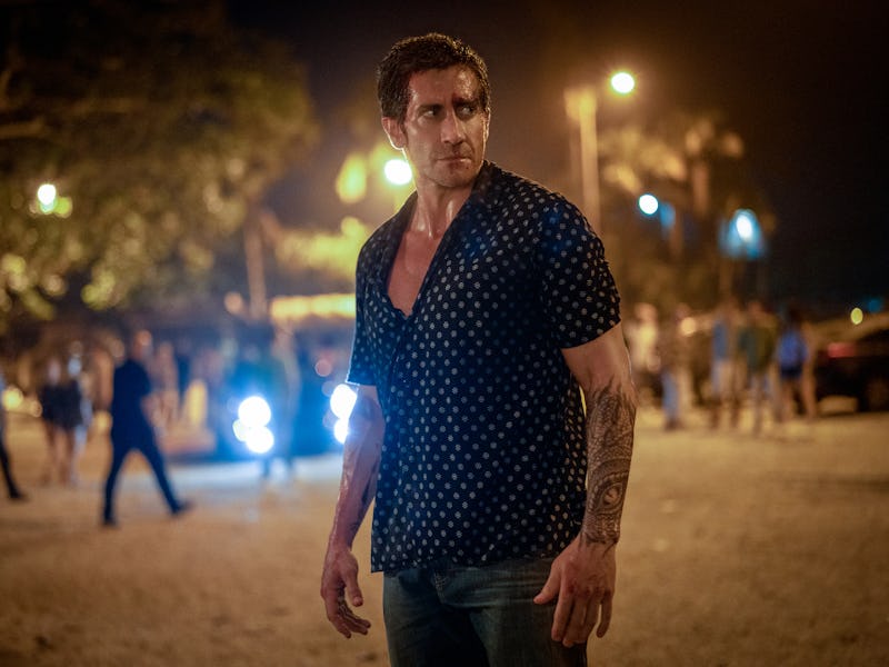 Man with a tattoo on his arm walking on a crowded beach at night, looking to the side with a serious...
