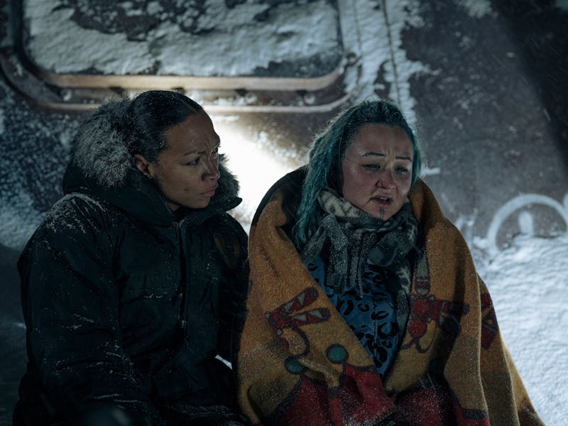 Two people huddled in winter clothing sit against a snowy background with a vehicle partially visibl...
