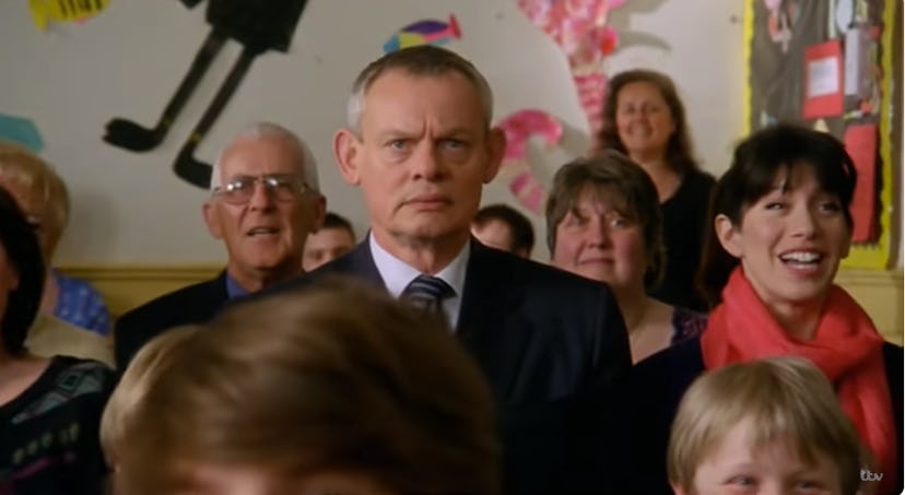 Doc Martin sits unsmiling in a crowd of happy people.