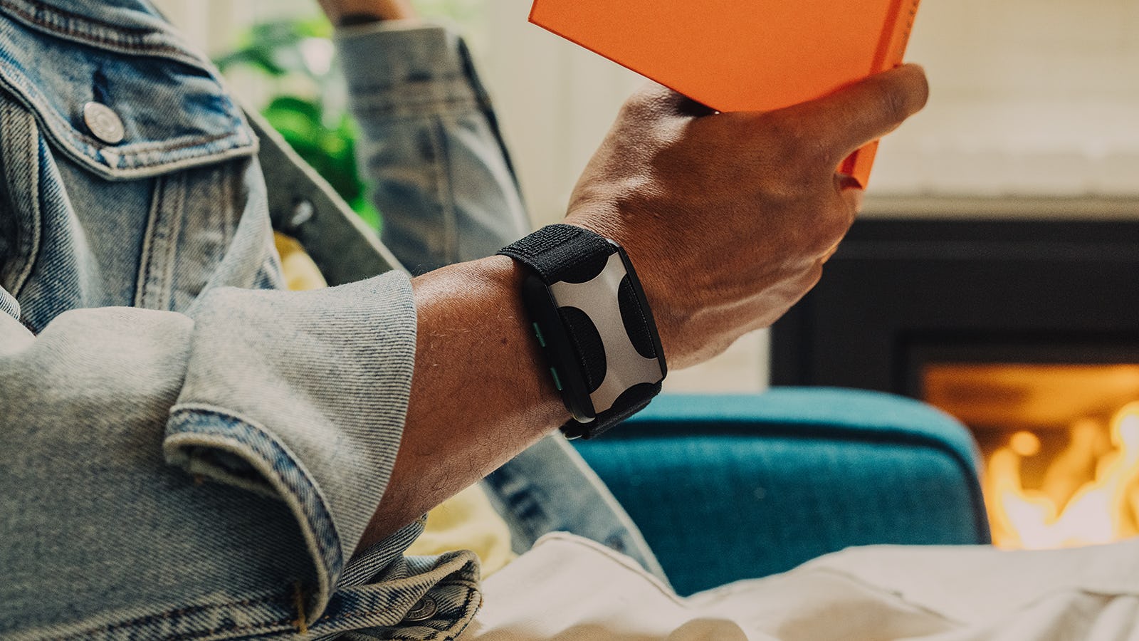 Person in denim jacket holding a book, wearing a black wearable device, with a cozy fireplace in the...