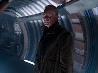 A man in a dark coat stands inside a dimly lit, futuristic corridor, looking intently to the side.