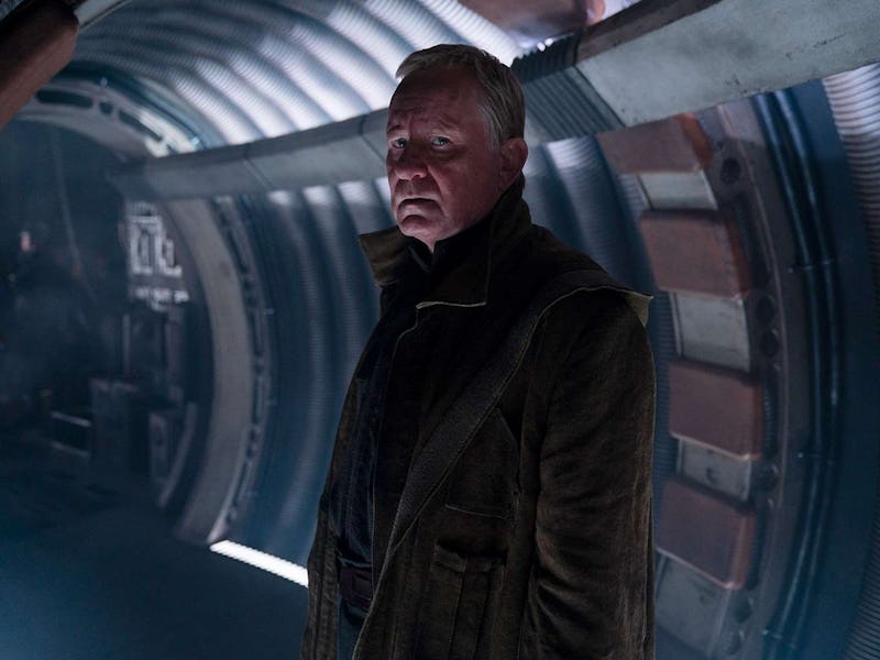 A man in a dark coat stands inside a dimly lit, futuristic corridor, looking intently to the side.