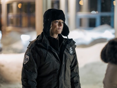 A person in a police uniform and fur hat stands outside at night with snow in the background, engage...