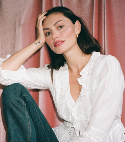 Woman sitting, wearing a white blouse and jeans, hand on head, against a pink backdrop.
