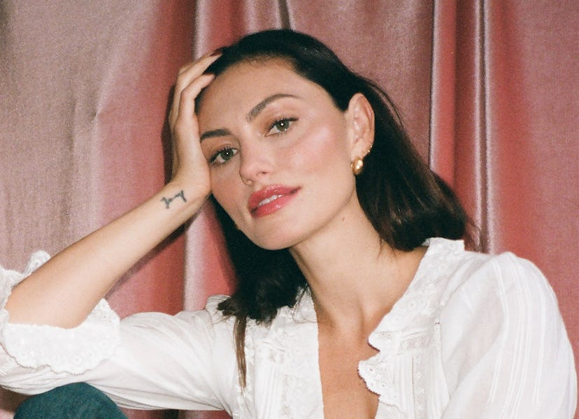Woman sitting, wearing a white blouse and jeans, hand on head, against a pink backdrop.