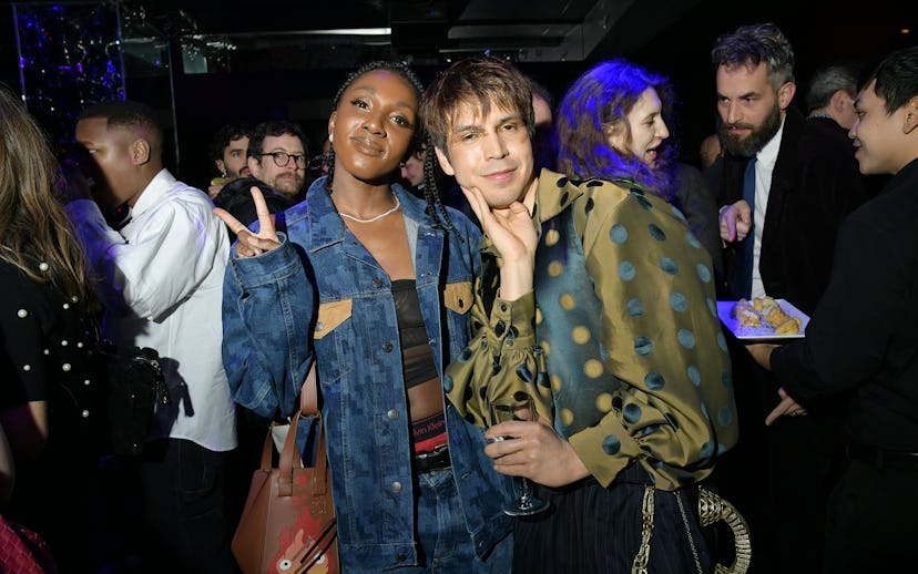 Two people posing together at a social event, surrounded by other guests in a dimly lit room.