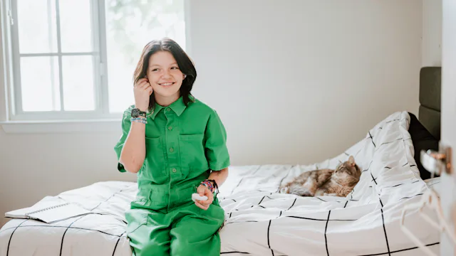 A nonbinary child sits on their bed with a cat.