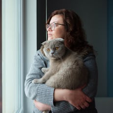 A woman solemnly looks out the window as she holds her cat in her arms.