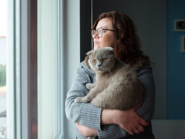A woman solemnly looks out the window as she holds her cat in her arms.