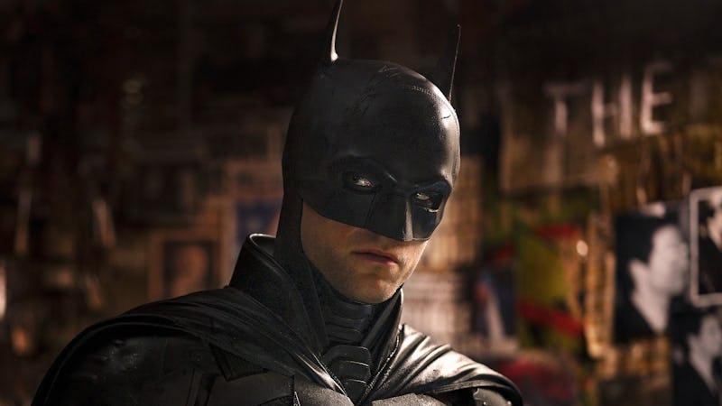 A person in a Batman costume with a cowl, looking intensely, against a background with posters and g...