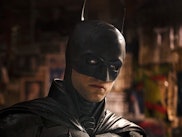 A person in a Batman costume with a cowl, looking intensely, against a background with posters and g...