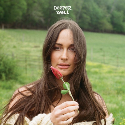 A woman holds a red flower to her face in a field, with text "DEEPER WELL" in the upper right.