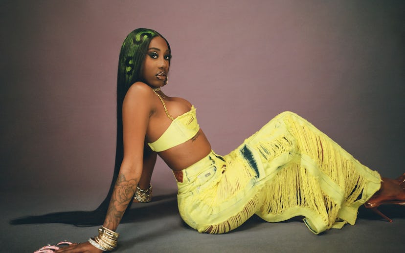 Woman with green highlighted hair in yellow crop top and pants posing on a purple background.