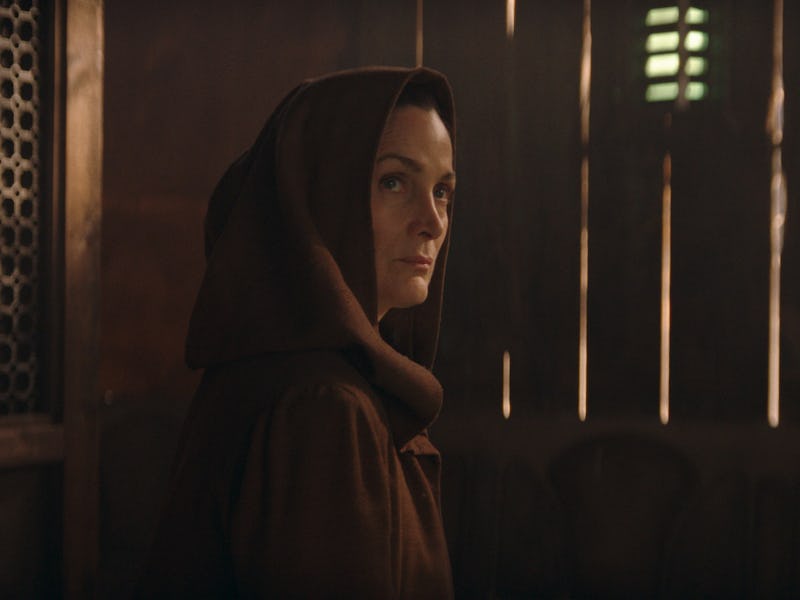 A woman in a hooded robe looks pensively to the side, with soft light filtering through vertical win...
