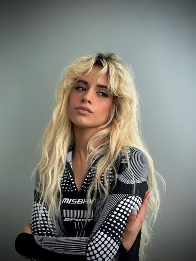 A person with tousled blonde hair, wearing a black and white patterned shirt, posing with a slight t...