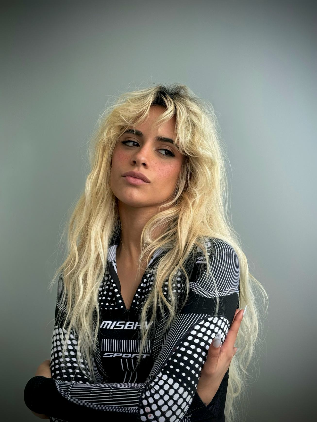 A person with tousled blonde hair, wearing a black and white patterned shirt, posing with a slight t...