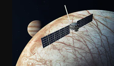 image of a moon with a cracked icy surface, with a satellite in the foreground and Jupiter in the ba...