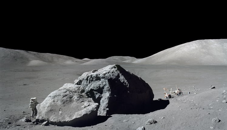 photo of an astronaut standing on a gray rocky surface, holding a science instrument, with a gold-fo...