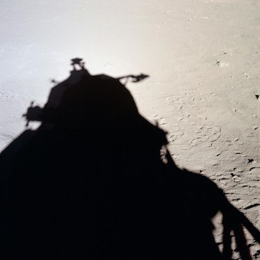 photo of a gray dusty surface with footprints, with the silhouette of a lunar lander part of shadowi...