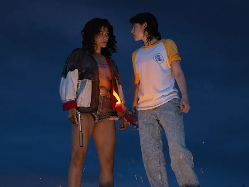 É noite. São duas mulheres, que vemos dos tornozelos pra cima. À esquerda, a personagem de Katy O'Brian, que é uma mulher branca de cabelos cacheados até os ombros, com um short jeans, um top e jaqueta, segurando um revolver com silenciador. Pelas pernas, é possível notar que ela é muito forte e musculosa. Ela está olhando para a personagem de Kristen Stewart, que é uma mulher branca e magra, cabelo liso um pouco mais curto e repicado. Ela está com uma calça jeans e uma camiseta comum. Em suas mãos, uma garrafa com um pano pegando fogo enfiado. As duas estão se olhando. | Cena do filme "O Amor Sangra"