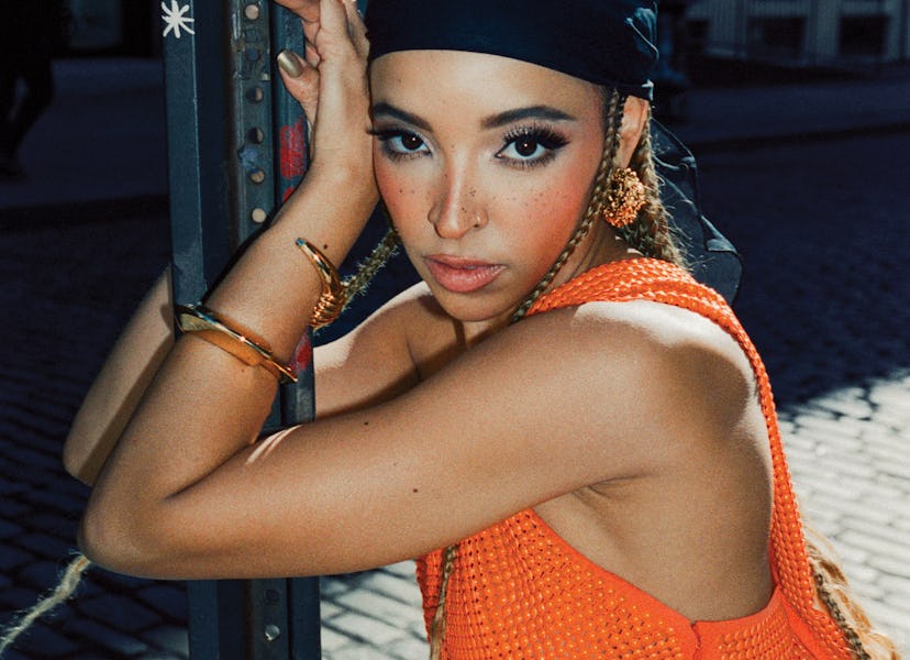 Woman with braided hair wearing a headscarf and orange top leans on a street pole.